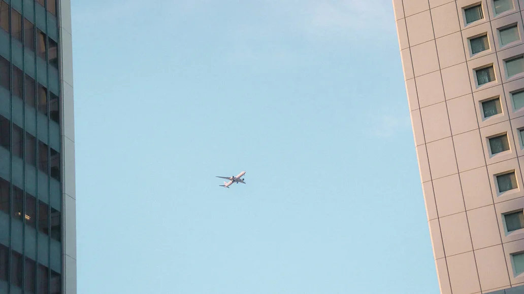 An airplane captured between two high-rise buildings under a pale blue sky.